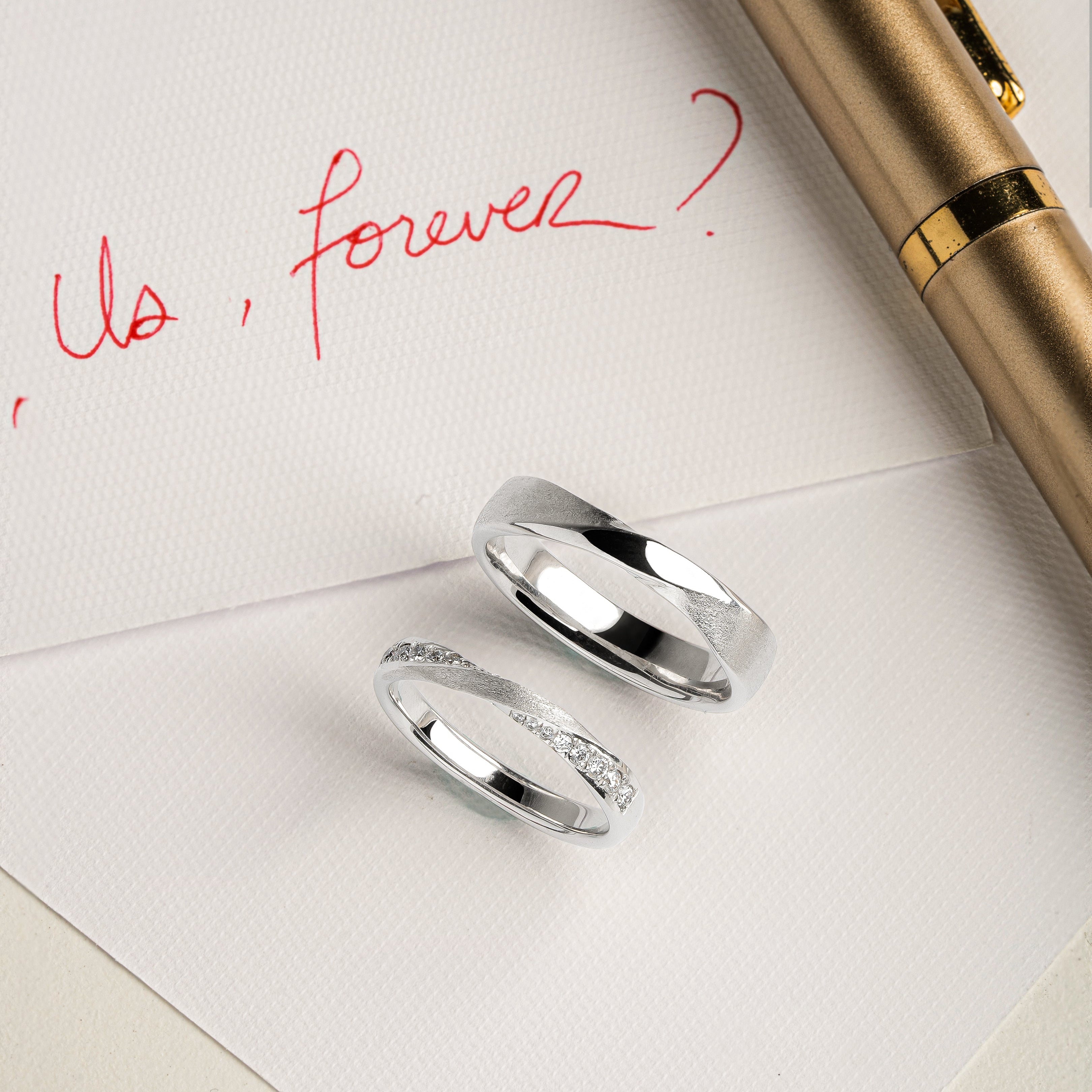 Two white Gold and Diamond Pave rings on a piece of paper with 'Us, forever?' written in red ink, accompanied by a gold pen.