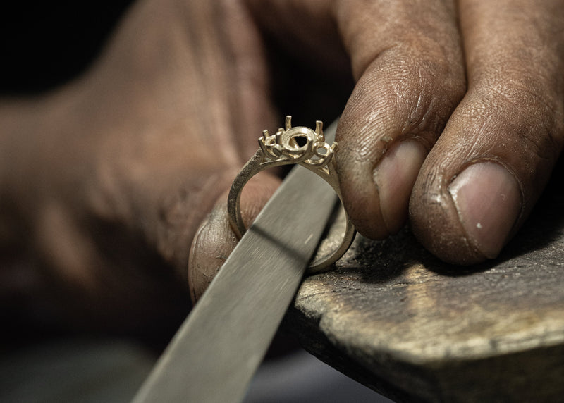 A close-up of a jeweler's hands holding a gold ring on a metal anvil, with a hammer resting nearby.